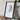 a framed artwork depicting a black and white abstract design, placed on a wooden surface next to a camera and some books.