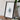 a framed artwork of a white flower on a wooden surface, with a camera and some books placed next to it.