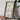 a framed map of a region, placed on a wooden surface next to a camera and some books.
