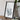 a framed map of a mountainous region, placed on a wooden surface next to a camera and some books.