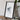 a framed artwork of a black and white abstract design, placed on a wooden surface next to a camera and some books.