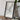 A framed map of the United States is displayed on a wooden table, accompanied by a camera, a book, and a plant.