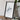 a framed artwork of a white, abstract shape on a wooden surface, with a camera and some books placed next to it.