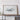 a large framed artwork depicting a landscape scene, placed on a wooden shelf against a white wall.