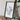 a framed artwork depicting a white, abstract design on a wooden surface, with a camera and books placed nearby.