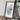 a framed artwork depicting a white, textured surface, placed on a wooden surface next to a camera and some books.