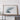 a large framed artwork depicting a landscape scene, placed on a wooden shelf against a white wall.