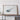 a large framed artwork depicting a stylized, abstract representation of a wave or shape, displayed on a wall above a wooden table with a camera and other items.