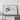 a large framed artwork depicting a mountainous landscape, placed on a wooden shelf against a white wall.
