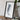 a framed artwork of a landscape, placed on a wooden surface next to a camera and some books.