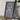 a framed map of the world, placed on a wooden table next to a camera and some books.