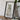 a framed map of the state of Alaska, with a camera and some books placed on a wooden table in front of it.