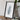 a framed artwork of a grayish-blue shape, placed on a wooden surface next to a camera and some books.