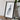 a framed artwork of a landscape, placed on a wooden surface next to a camera and some books.