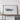 a large white framed artwork depicting a landscape with a blue and white color scheme, placed on a wooden shelf against a white wall.