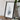 a framed artwork of a landscape, placed on a wooden surface next to a camera and some books.
