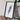 a framed artwork of a mountain range, placed on a wooden surface next to a camera and some books.