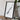 a framed artwork of a black and white abstract design, placed on a wooden surface next to a camera and some books.