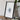 a framed artwork of a leaf-like design on a white background, placed on a wooden surface next to a camera and some books.