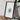 a framed artwork of a white flower on a wooden surface, with a camera and a book placed next to it.