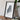a framed artwork of a mountain range, placed on a wooden surface next to a camera and some books.